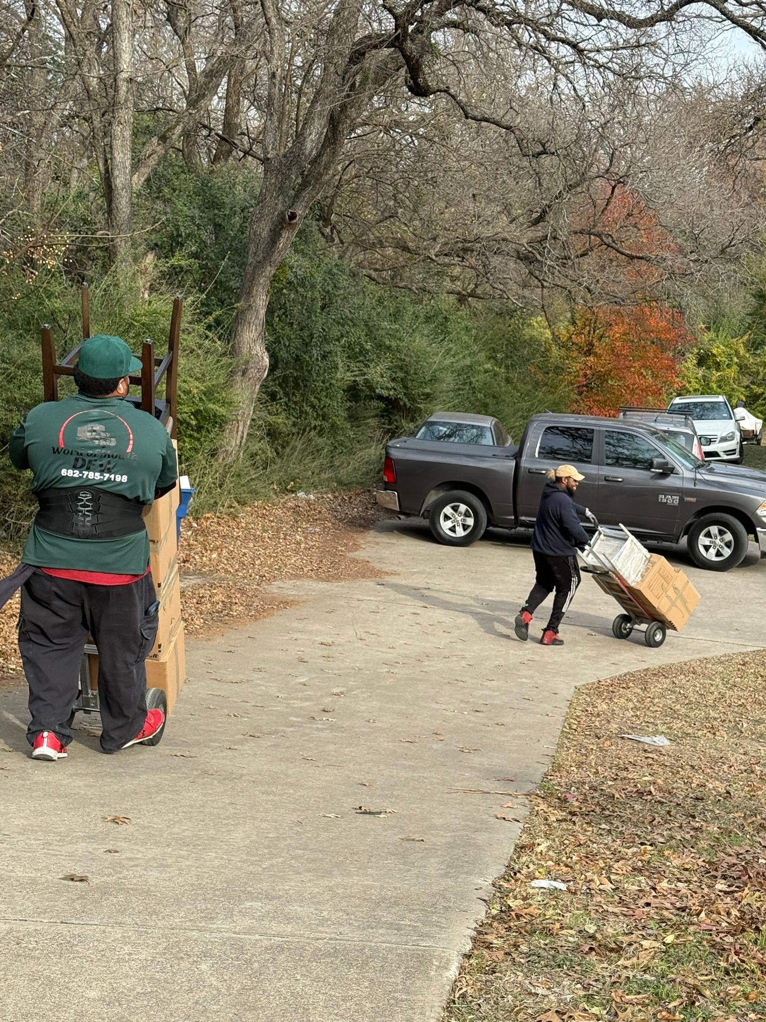 Two people moving boxes and furniture next to trucks on a driveway near trees.