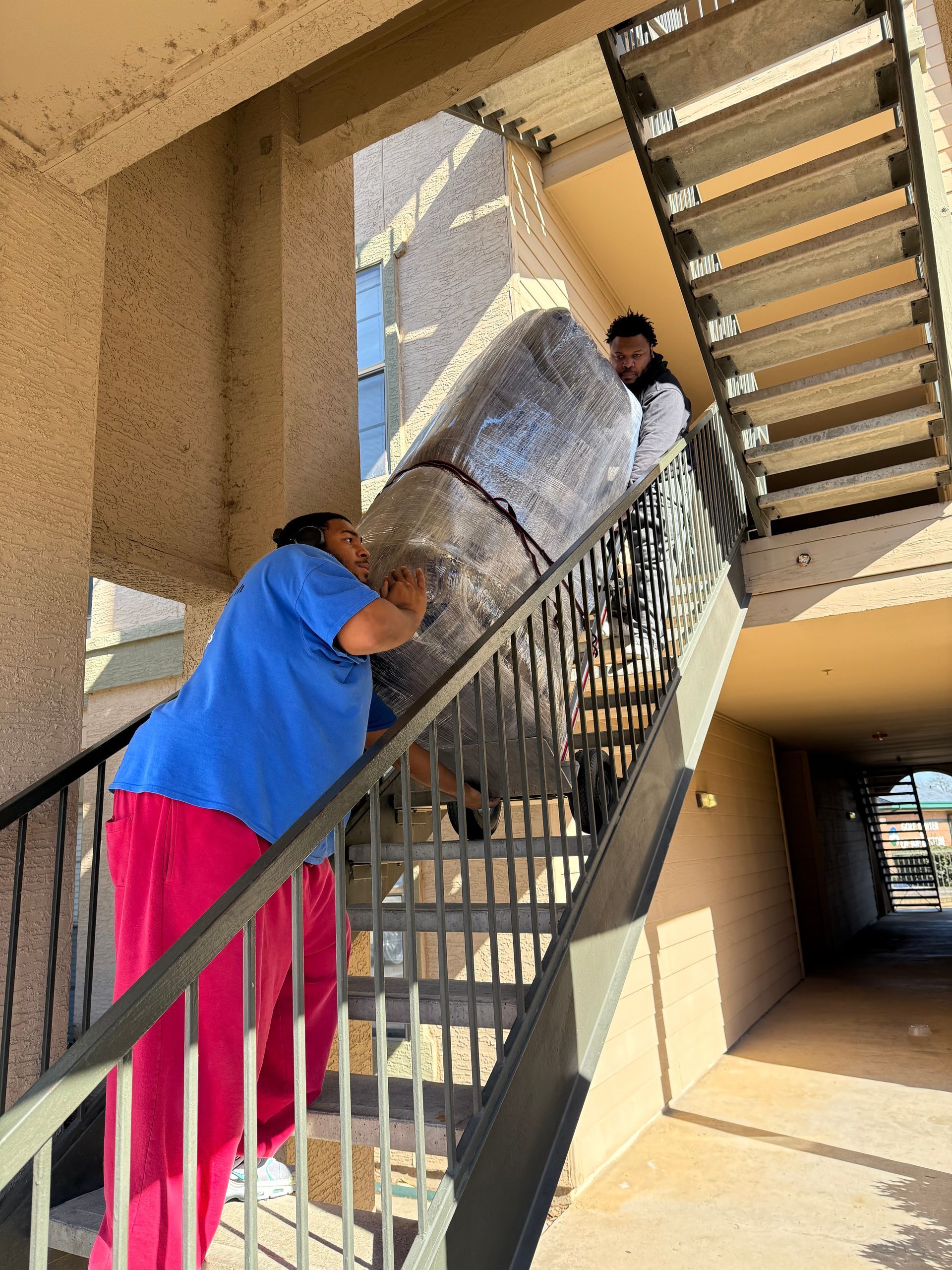 Two people carry a large silver panel up an outdoor stairwell under a building.