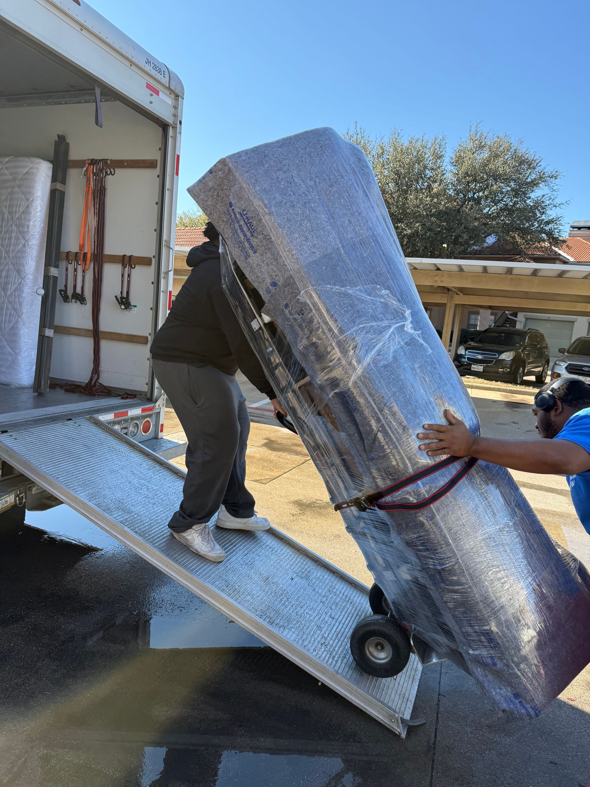 Two movers unload a large wrapped appliance from a truck ramp outdoors.