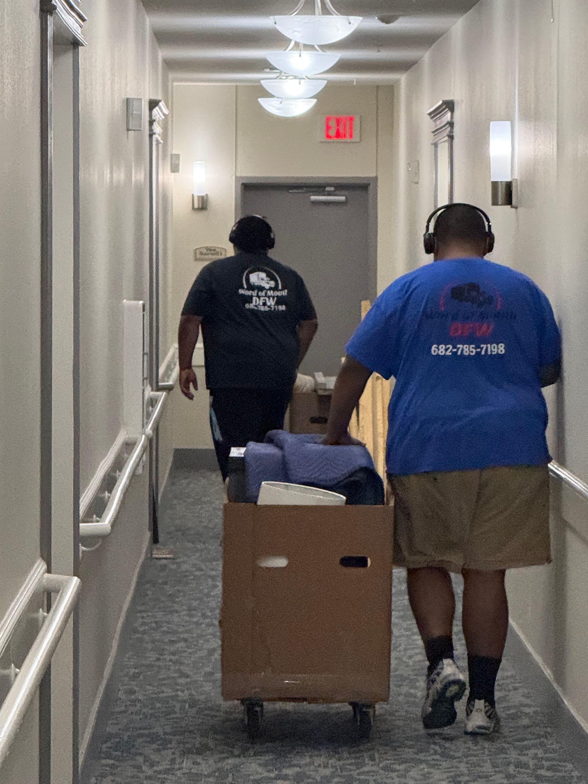Two people push a cart with a box down a narrow hallway toward a closed door.