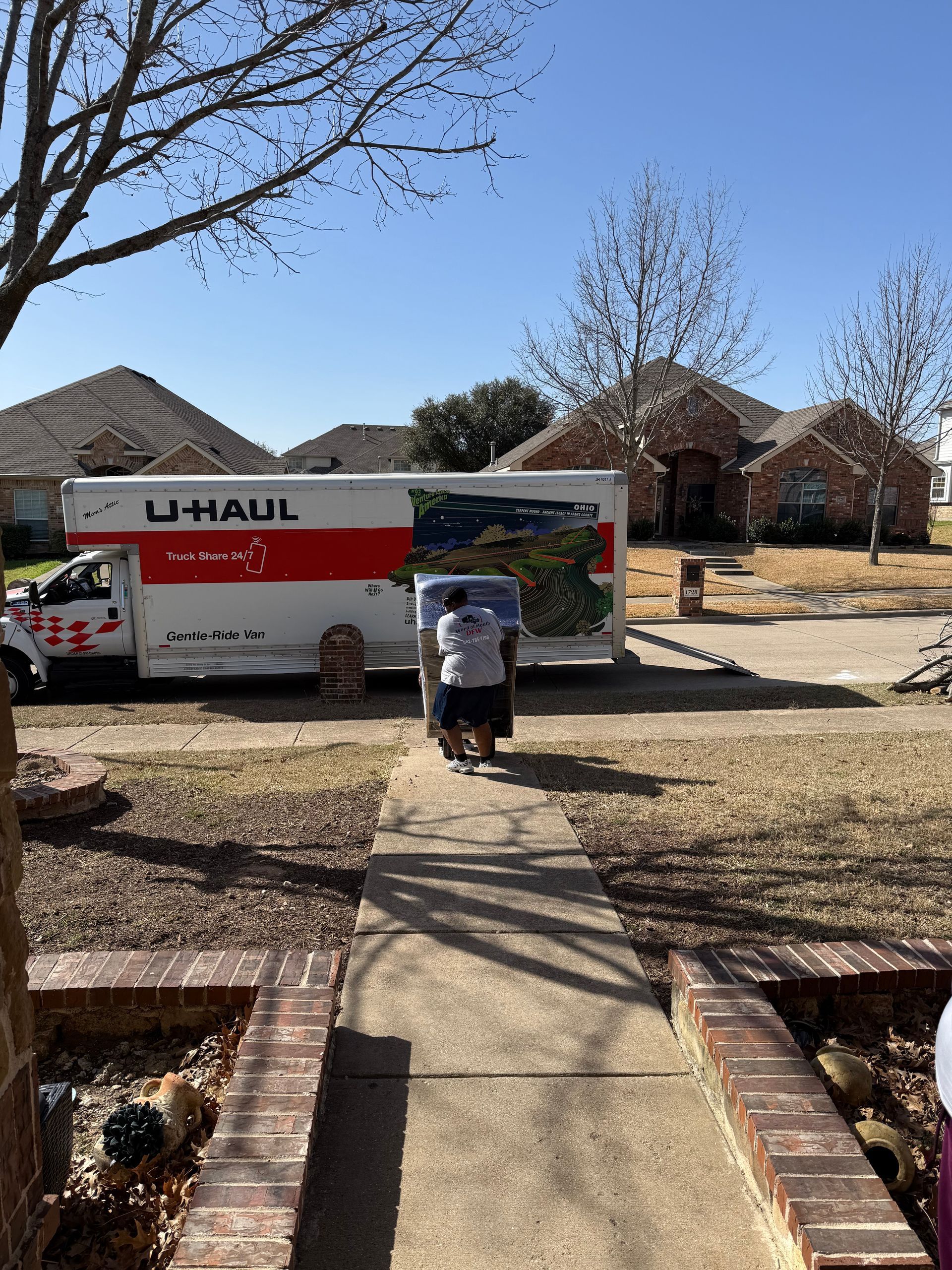 U-Haul truck parked in a suburban driveway, with a person carrying boxes toward it under a clear blue sky