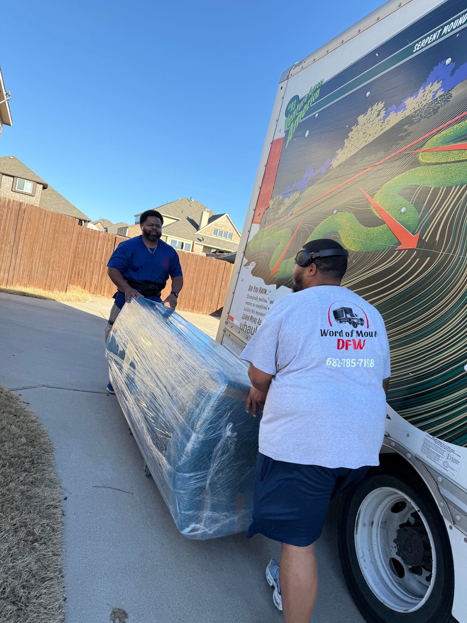 Two workers unload a wrapped mattress from a truck in a driveway.