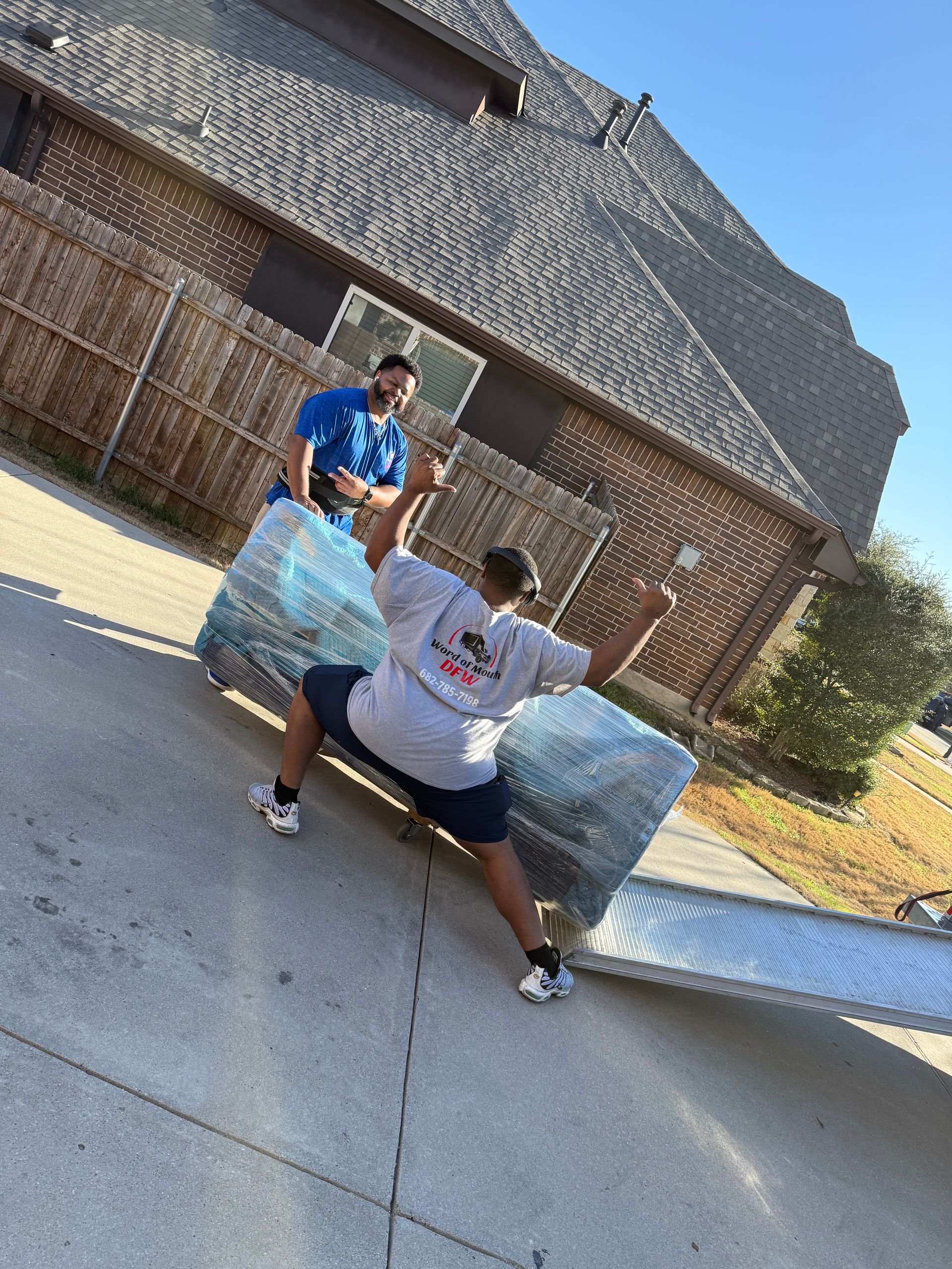 Two people pushing a large blue-wrapped object in a driveway beside a brick house.