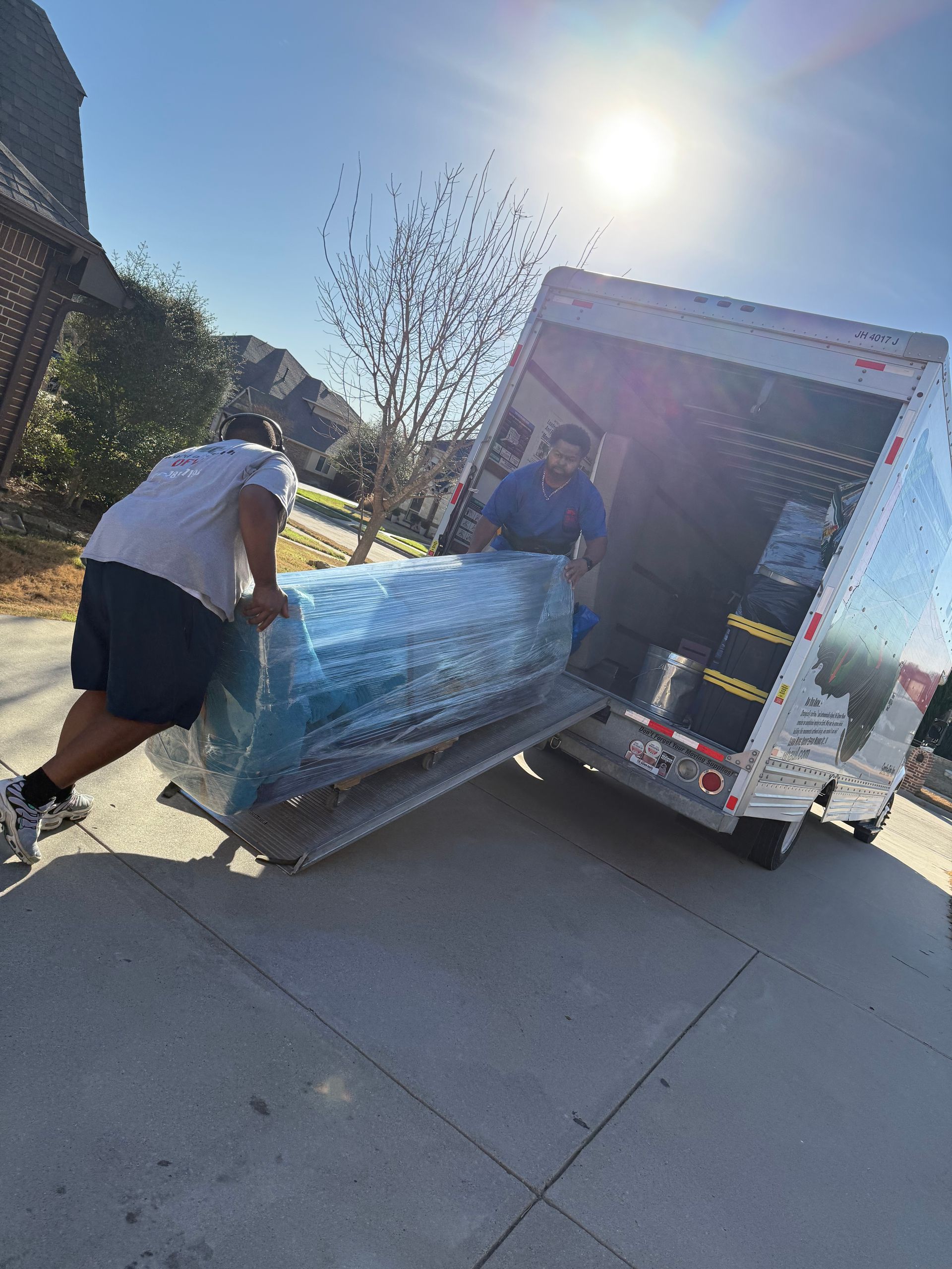Two people loading a wrapped couch into a moving truck in a sunny driveway