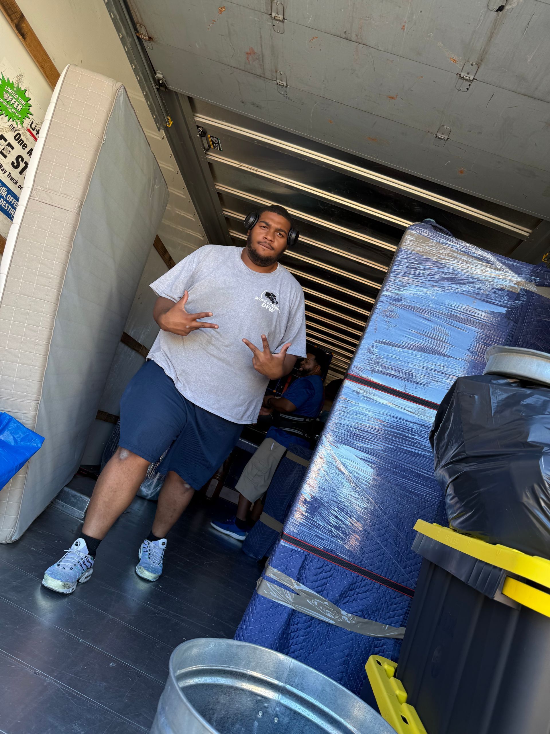 Man standing in a truck or storage area beside stacked blue-wrapped boxes and yellow-handled equipment.