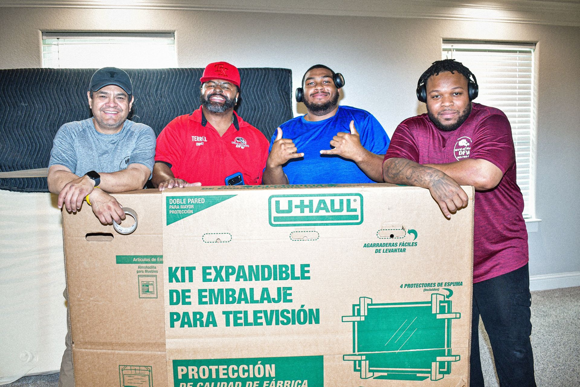 Four people pose behind a large U-Haul moving box indoors, smiling at the camera.