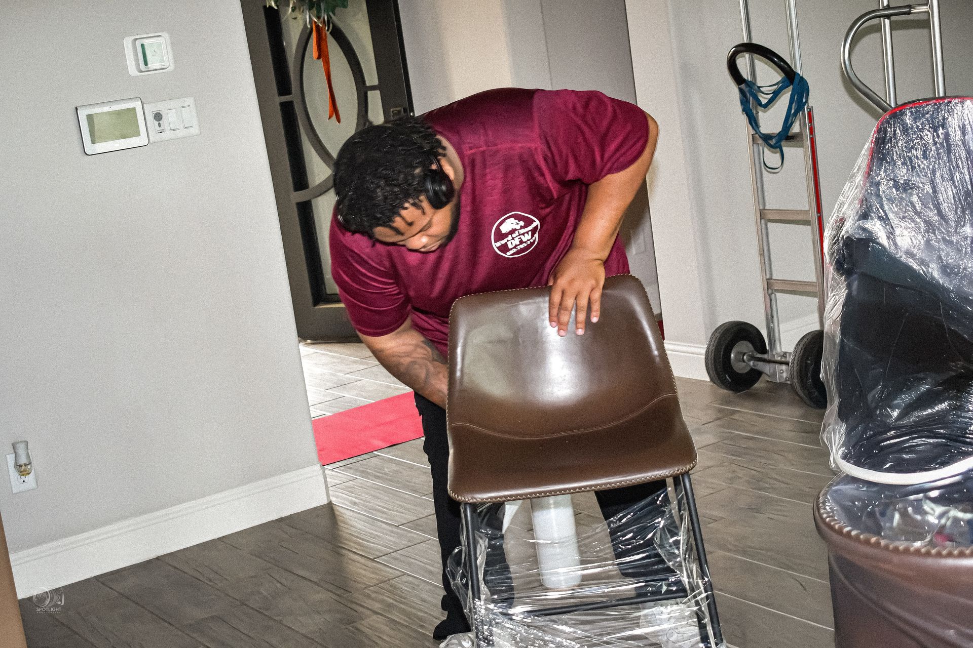 Person adjusting a brown chair wrapped in plastic in a room with stacked furniture