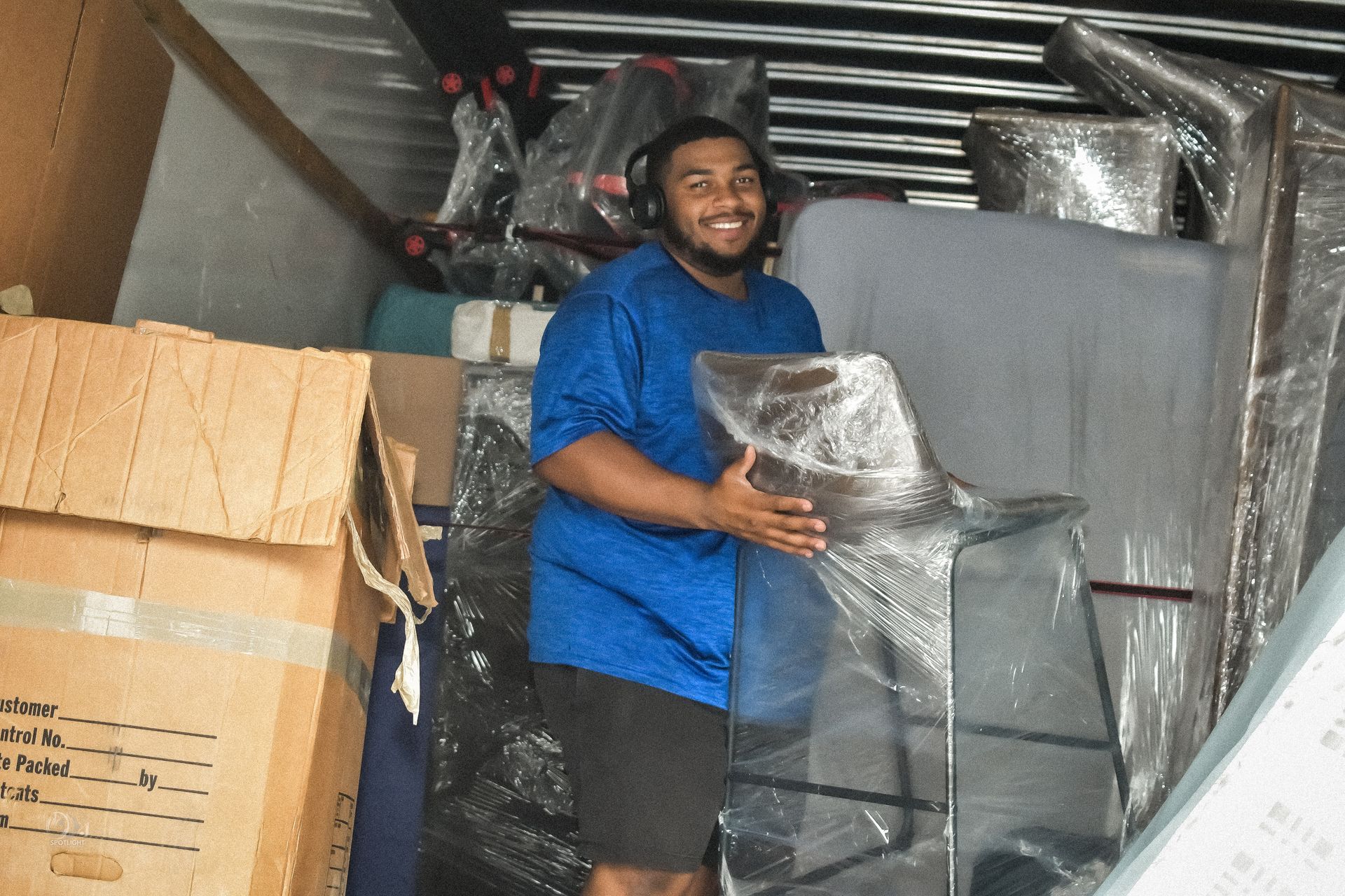 Man smiling while holding a wrapped chair in a storage area surrounded by packed furniture and boxes