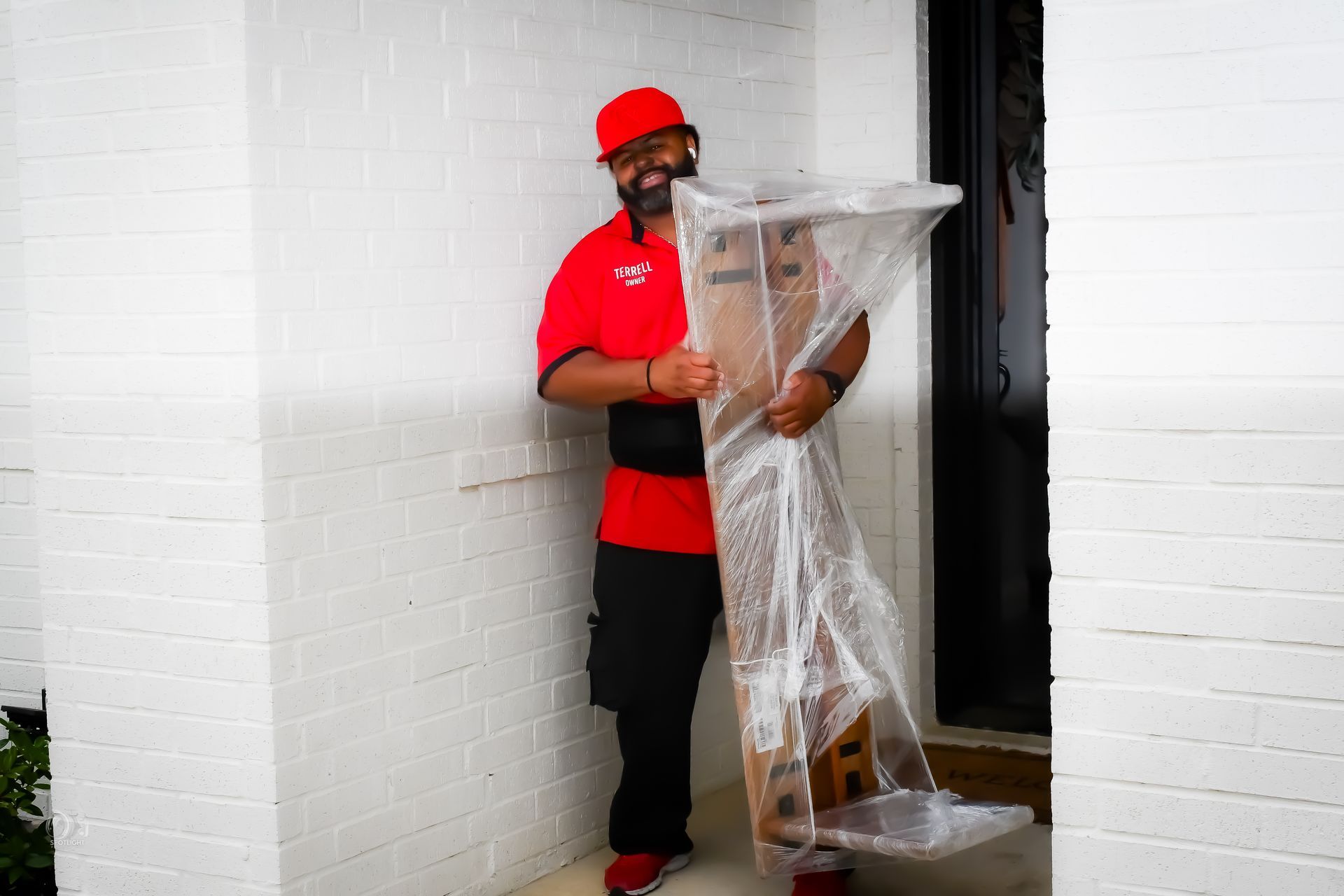 Man in red cap and shirt holding a clear plastic cover in a doorway, standing by a white brick wall
