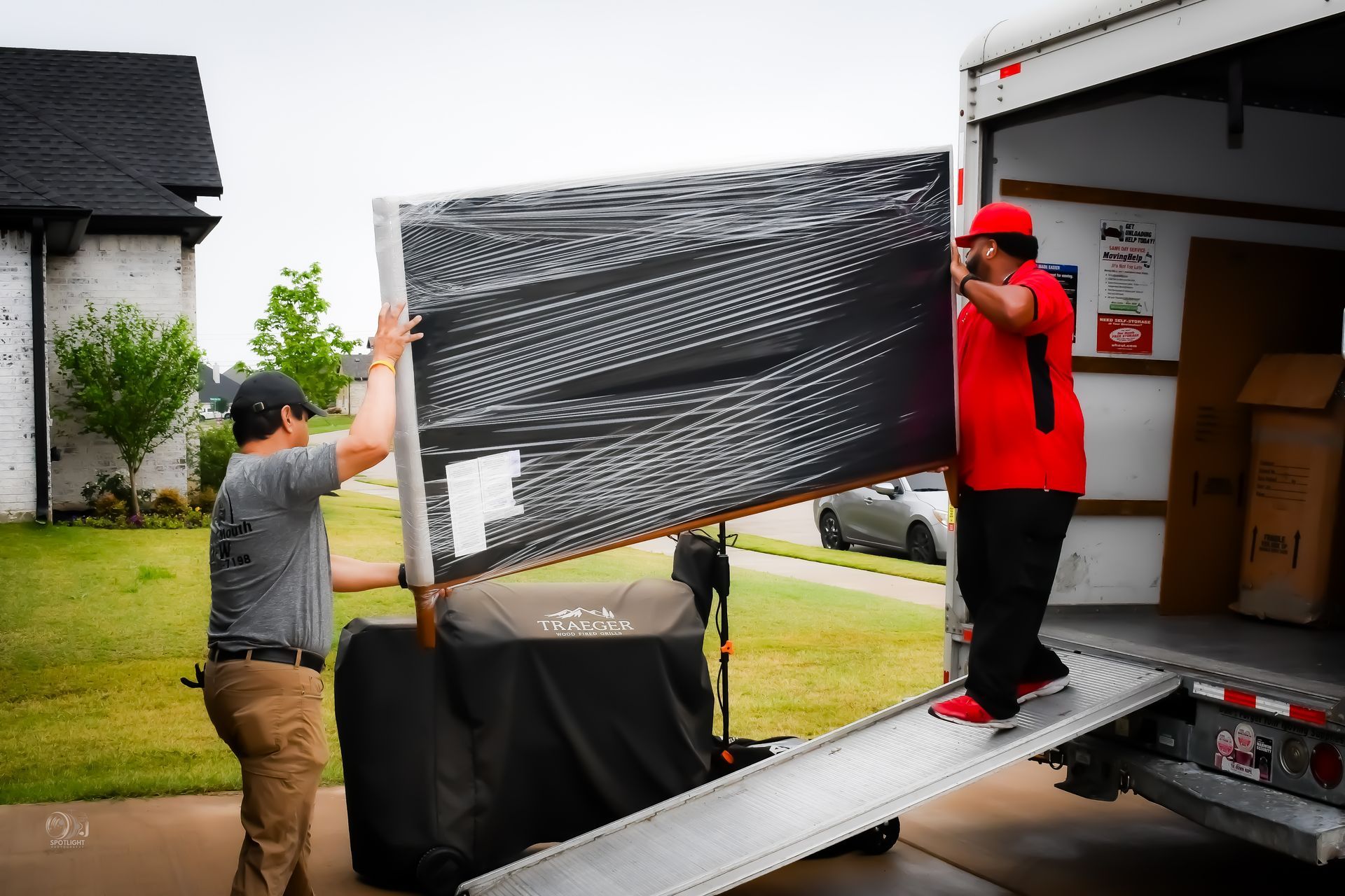 Two men loading a large wrapped TV into a moving truck from a home driveway.