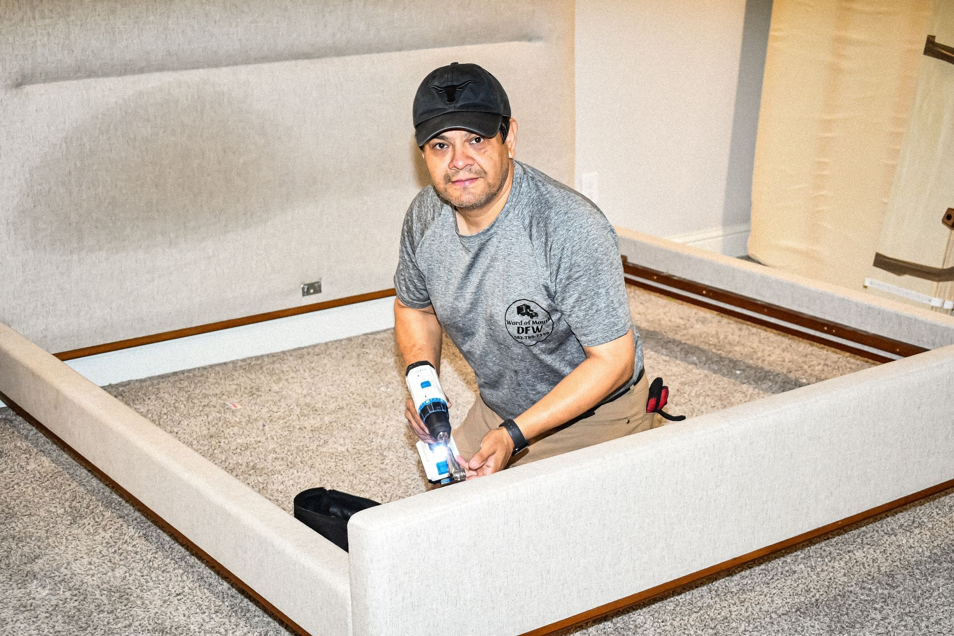 Person assembling a large upholstered bed frame in a bedroom, using a blue tool.