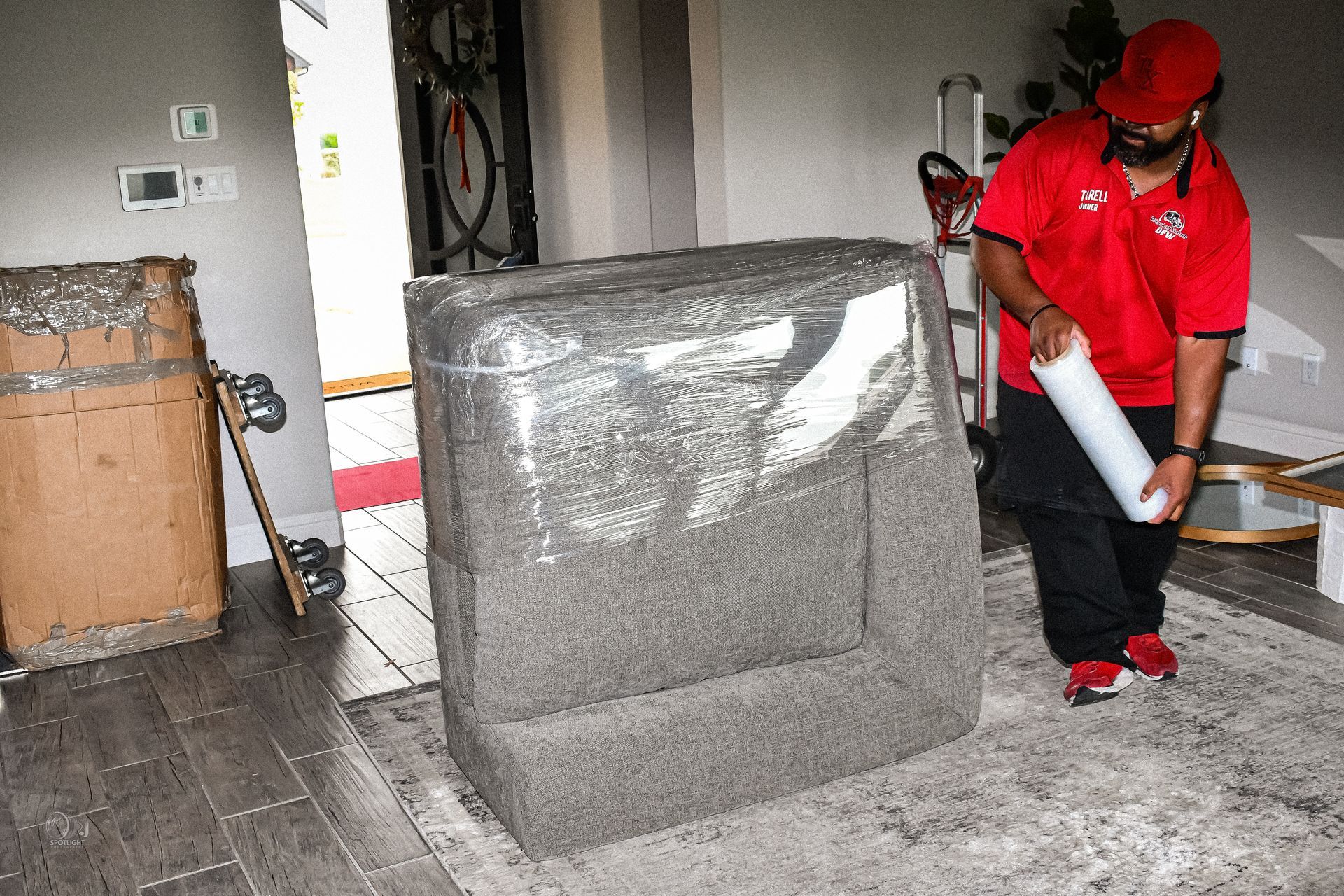 Worker in red uniform cleaning a dusty room beside a large wrapped sofa