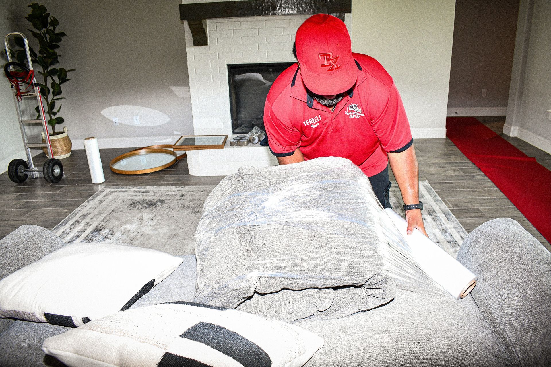 Person in a red shirt leaning over a large gray sofa in a bright living room.