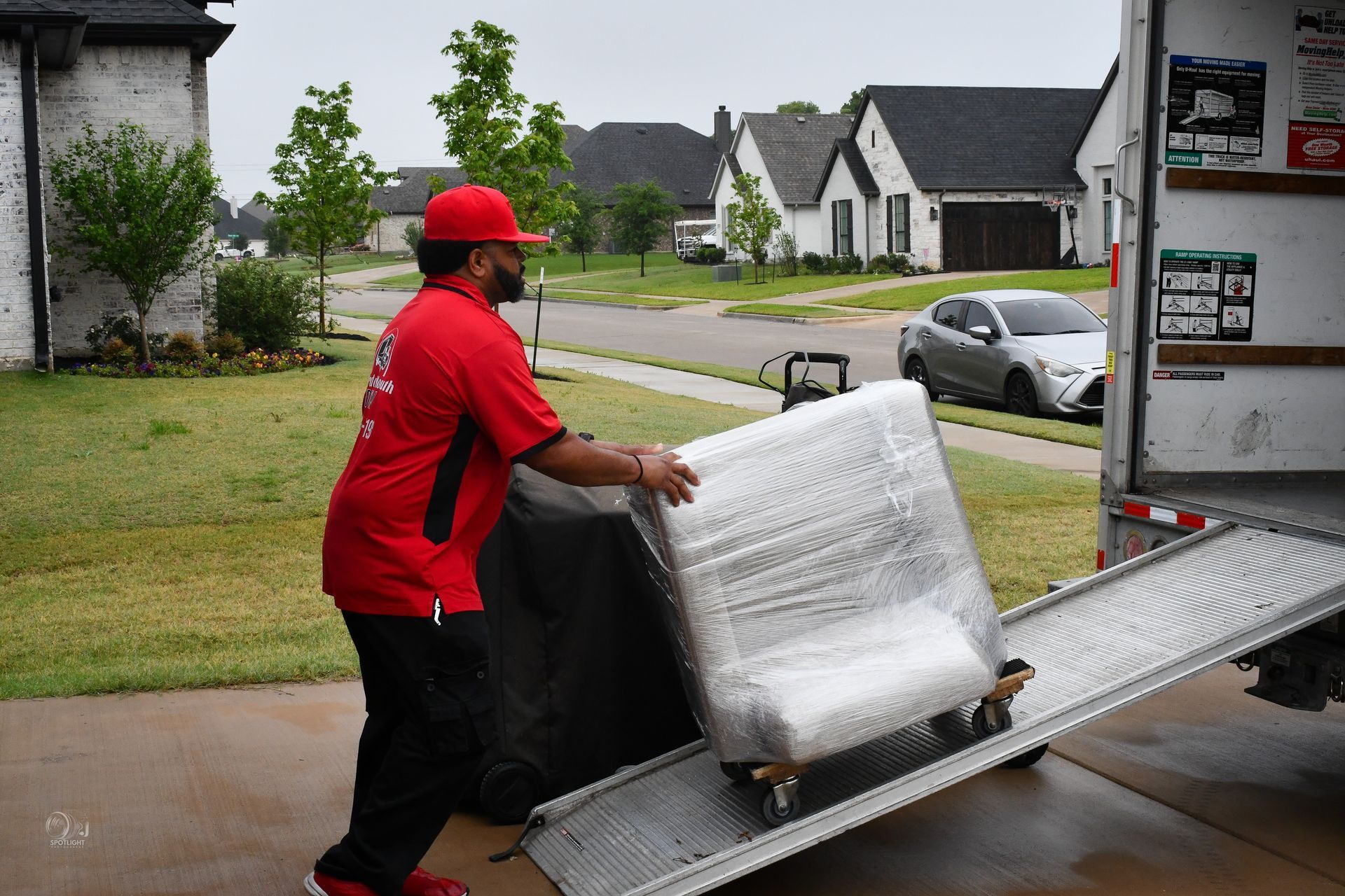Worker in red uniform pushing a wrapped appliance up a truck ramp in a suburban driveway