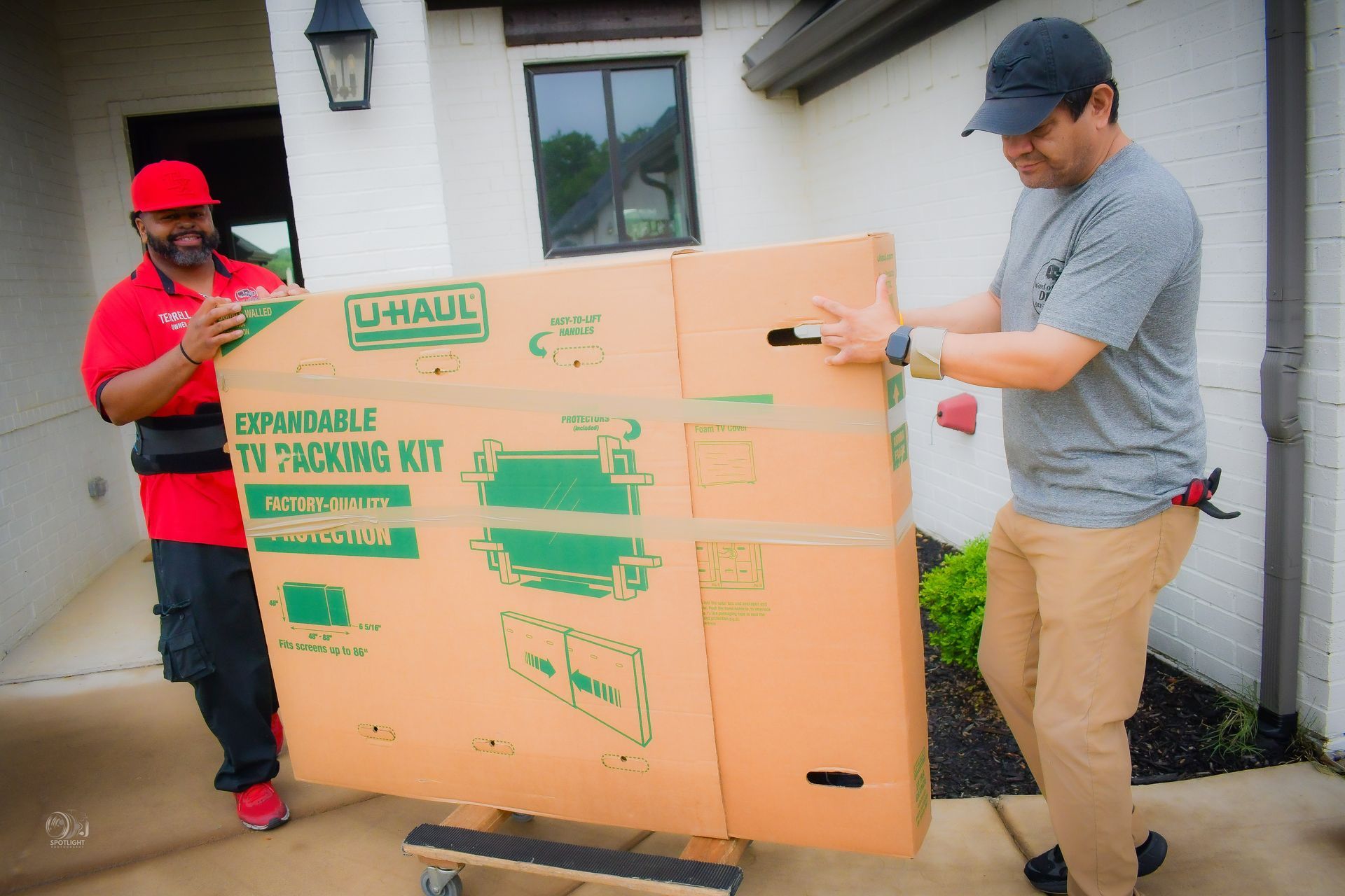 Two people moving a large cardboard box outside a house, one using a red hand truck.
