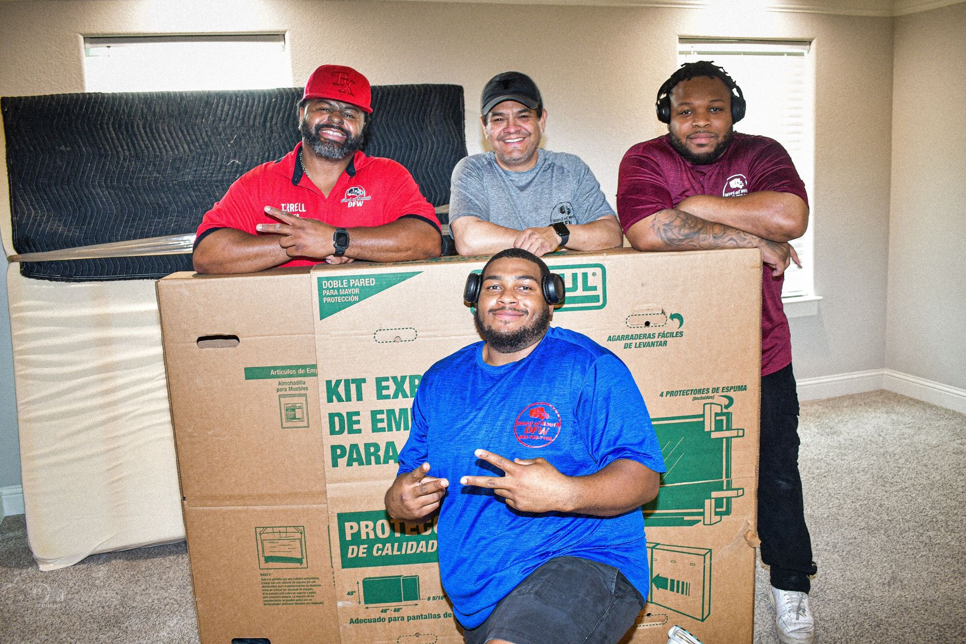 Four people posing with a large cardboard box indoors, three standing behind it and one kneeling in front.