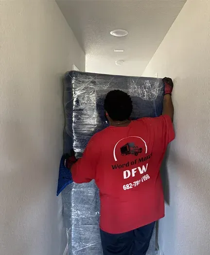A man in a red shirt is carrying a mattress in a hallway.