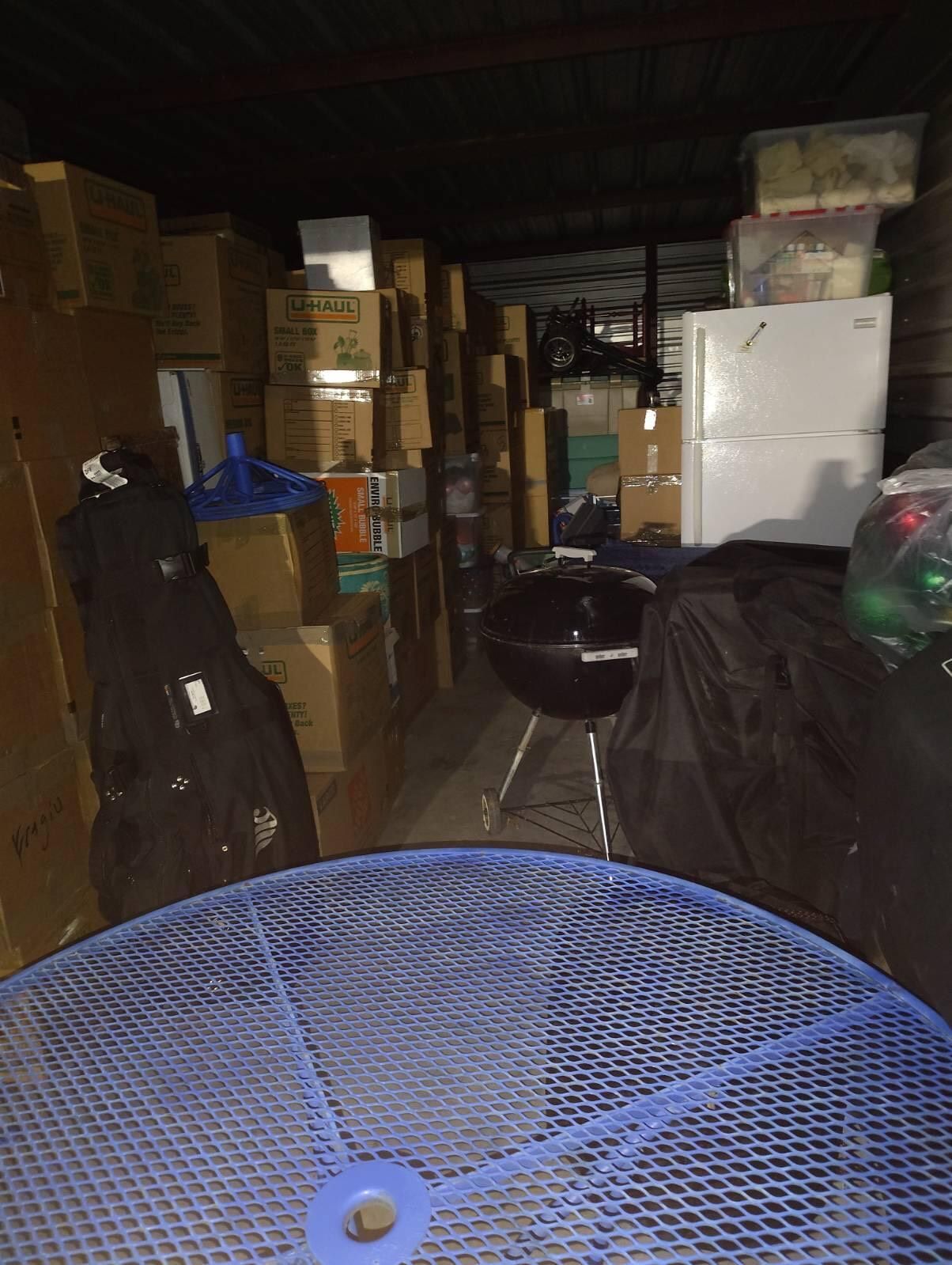 Storage unit, full of boxes and appliances, dark interior, blue table in foreground.