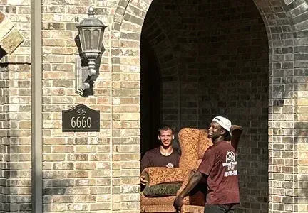 Two men are carrying a couch in front of a brick building.