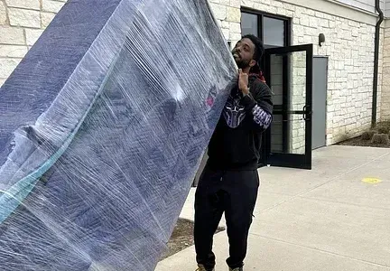 A man is carrying a large piece of furniture wrapped in plastic.
