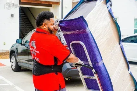 A man in a red shirt is carrying a mattress on a cart.