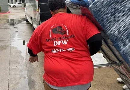 A man in a red shirt is carrying a mattress on his back.