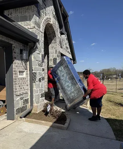 A man in a red shirt is carrying a refrigerator into a house.