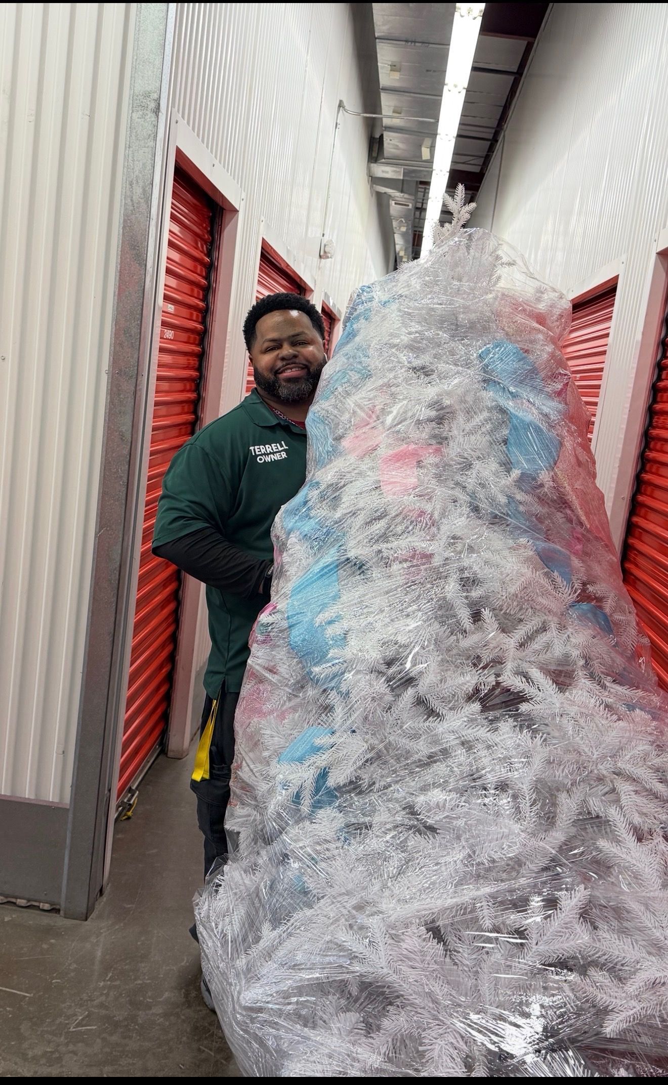 Person standing beside a large white and blue artificial Christmas tree in a storage hallway.