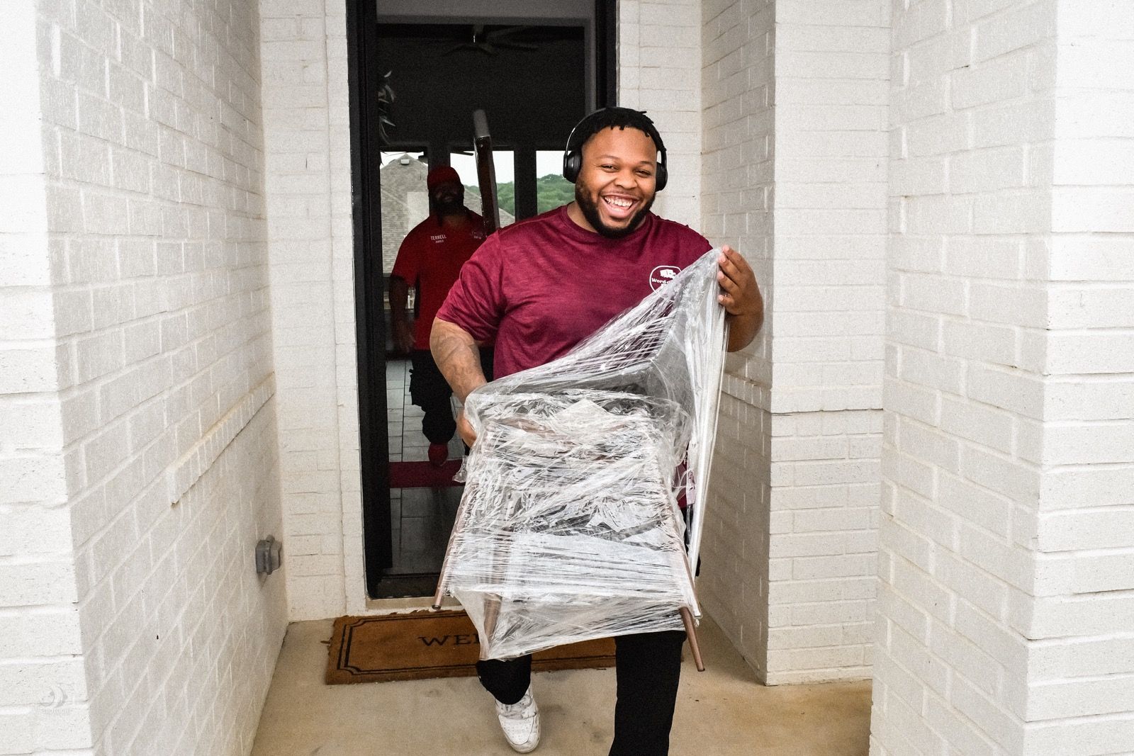 Smiling person in a maroon shirt carrying a plastic-wrapped package in a doorway
