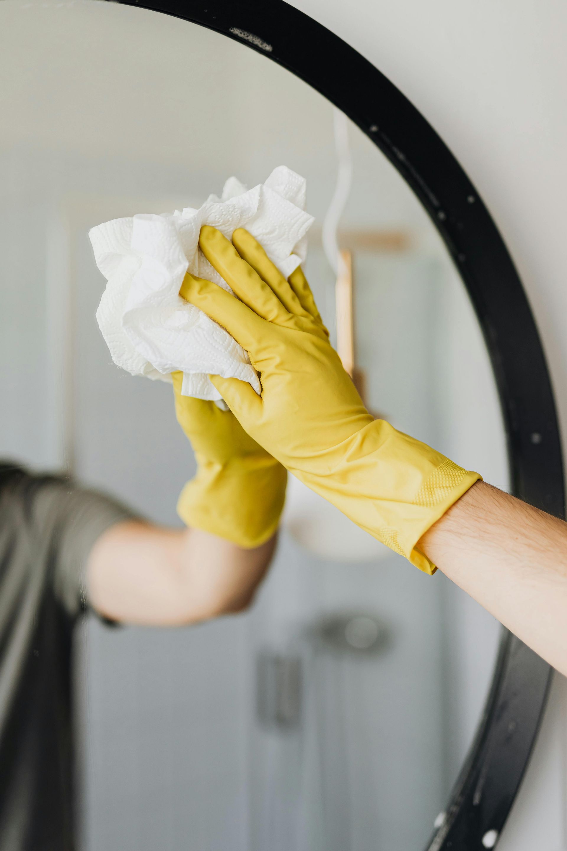 Person in yellow gloves cleaning a round mirror with a white cloth.