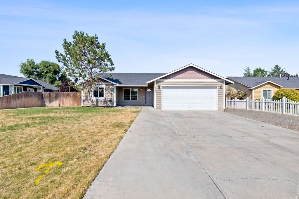 A single-story, light-colored suburban house with a white garage door, driveway, and dry grass under a blue sky.