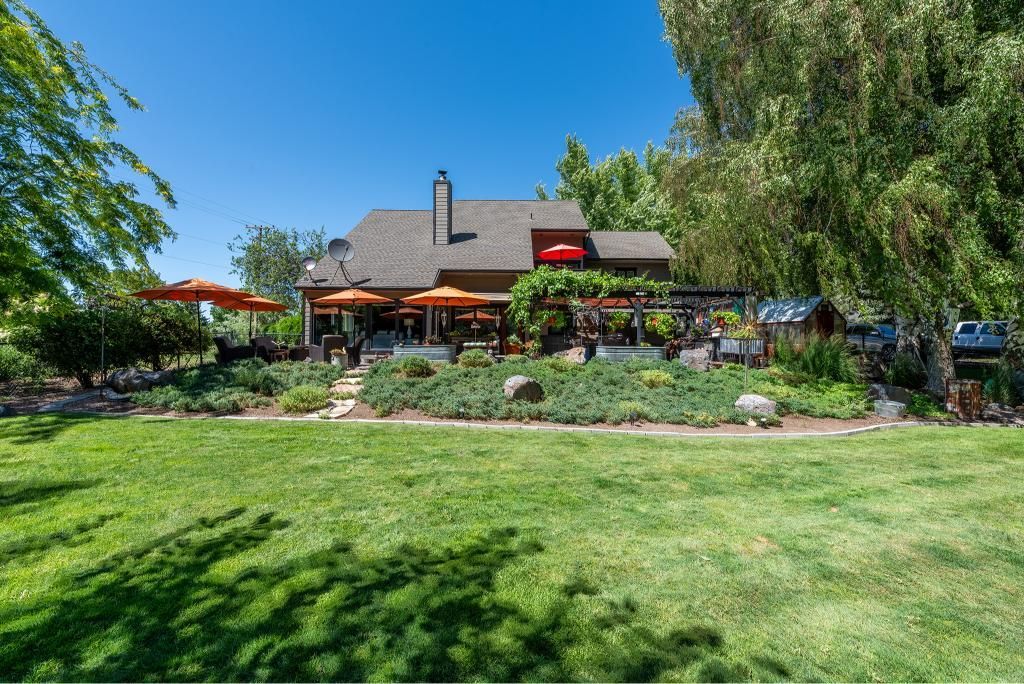 A sunny restaurant with a gray roof and outdoor patio seating under orange umbrellas, surrounded by lush green trees.