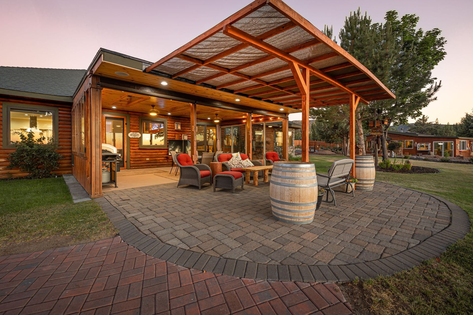 A cozy wooden patio with a pergola, outdoor seating, and wine barrels on a paved stone terrace at dusk.