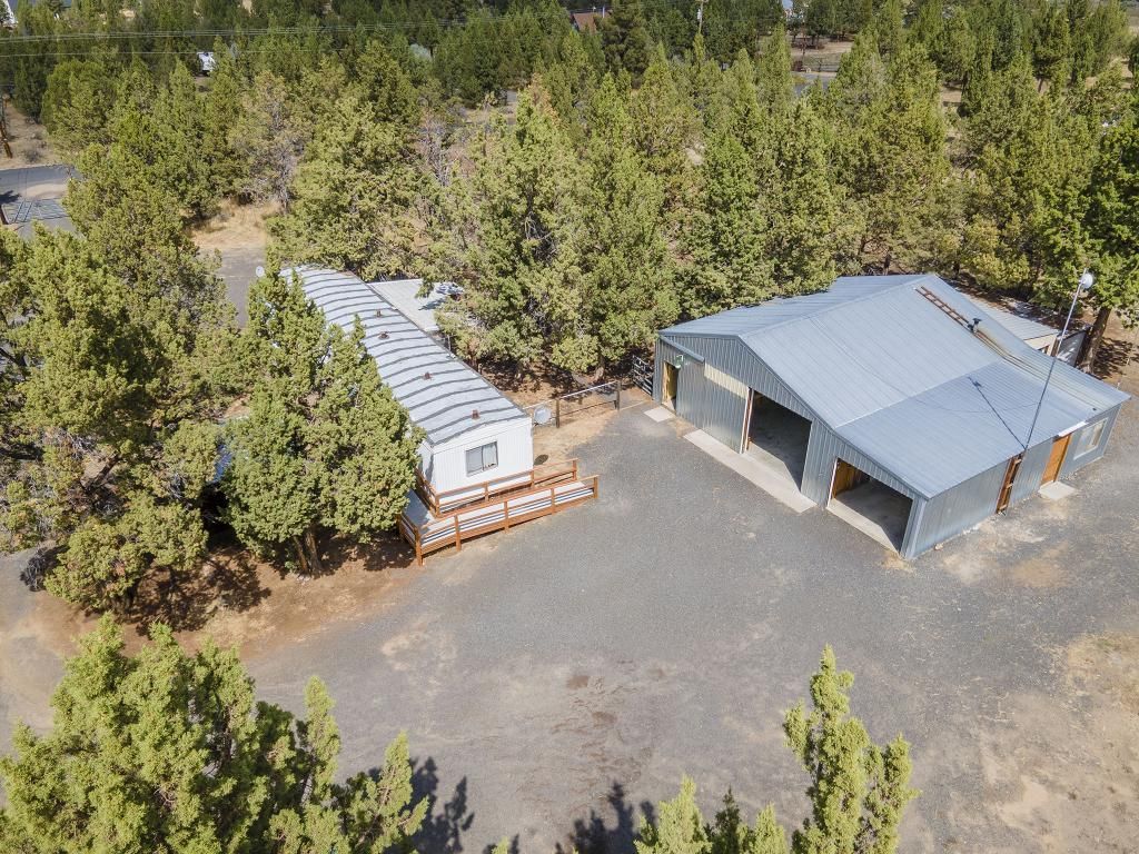 Aerial view of a white mobile home with a wooden deck next to a large, grey metal workshop on a gravel lot in a forest.