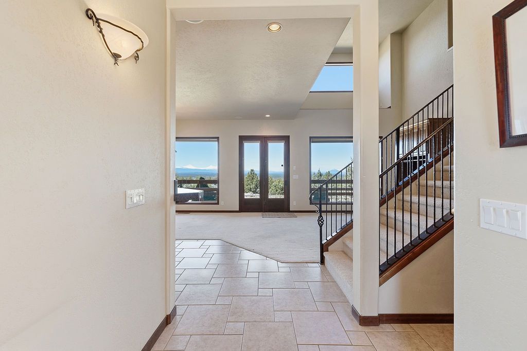 An interior foyer with tiled floors, a staircase with metal railings, and a view into a living area with large windows.