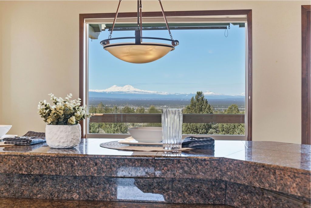A granite kitchen island with a vase of flowers faces a large window with a scenic view of trees and snow-capped mountains.