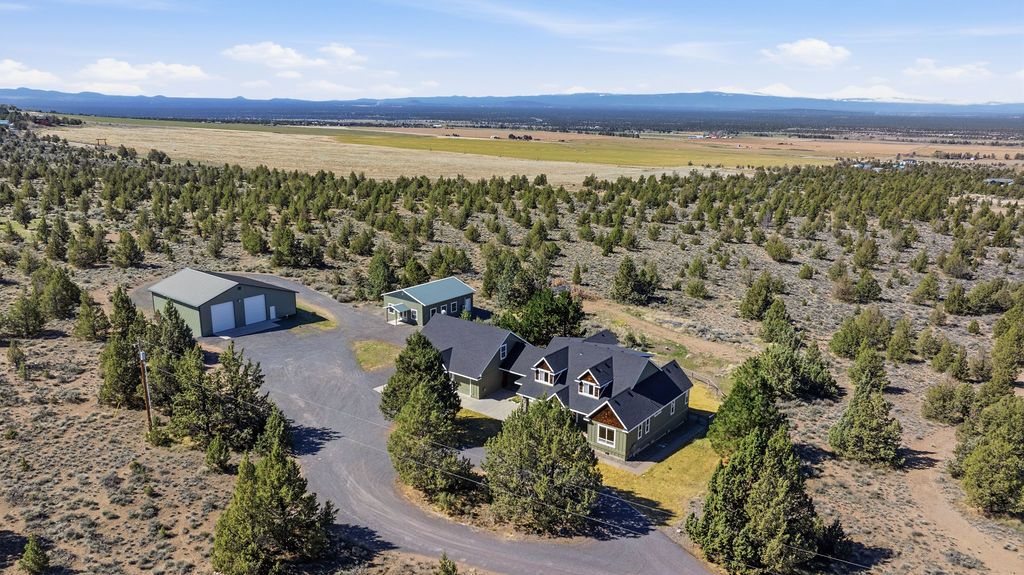 Aerial view of a house and detached garage surrounded by juniper trees and desert scrubland under a blue sky.
