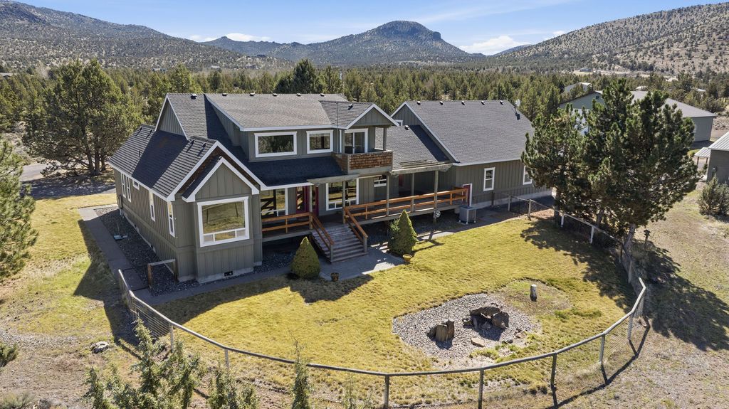 A gray two-story house with a wooden deck and fire pit, situated in a rural landscape with trees and distant hills.