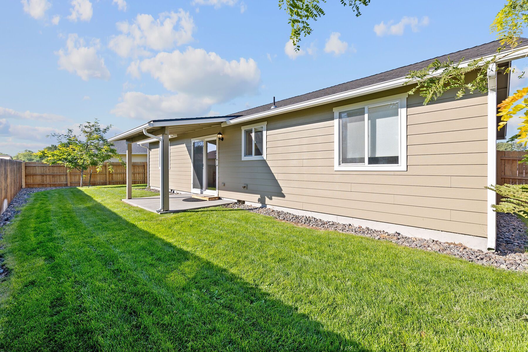 Back view of a tan, single-story house with a covered patio, window, and a neatly mown lawn under a clear blue sky.