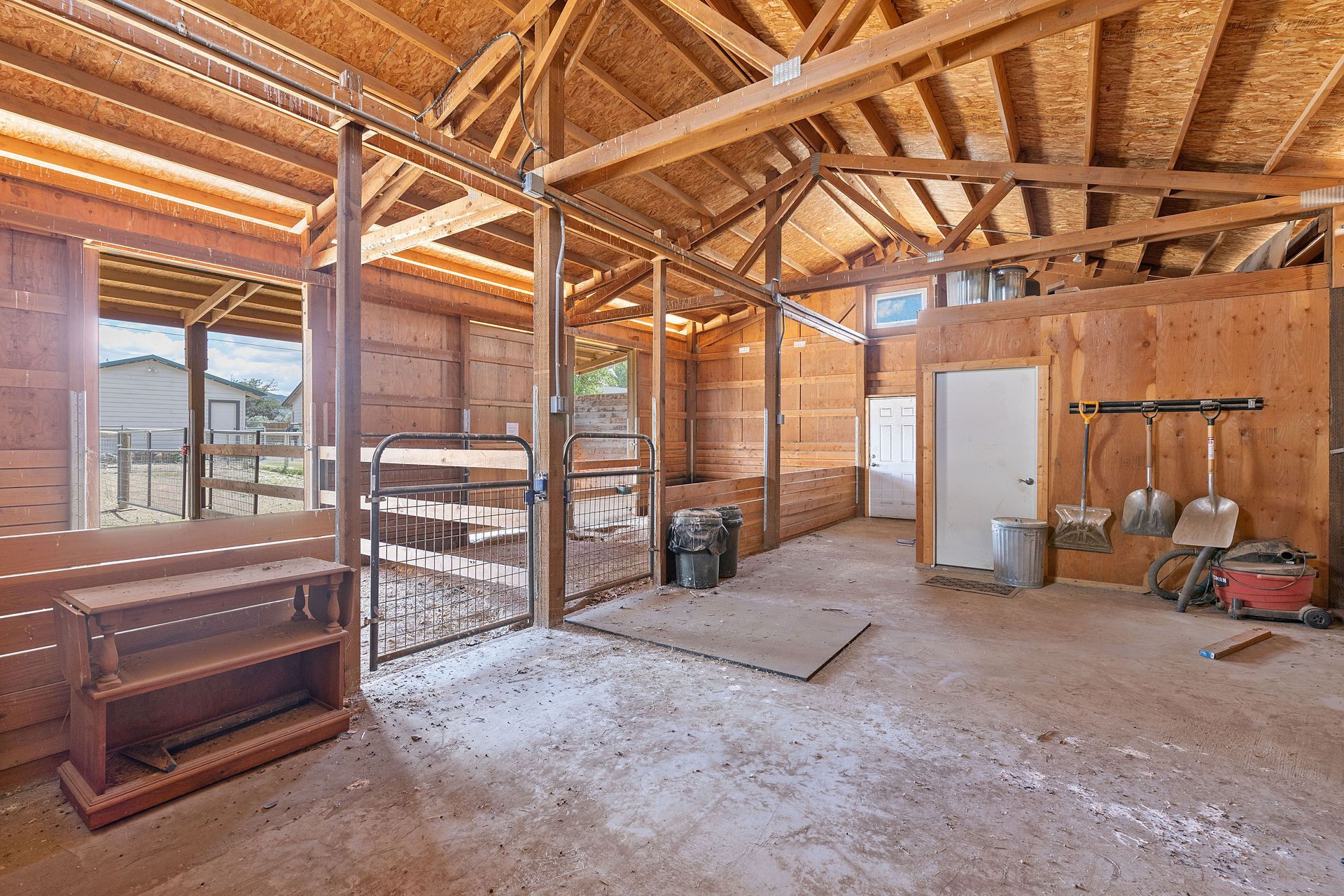 The interior of a wooden barn featuring multiple horse stalls, a concrete floor, and tools hanging on the wall.