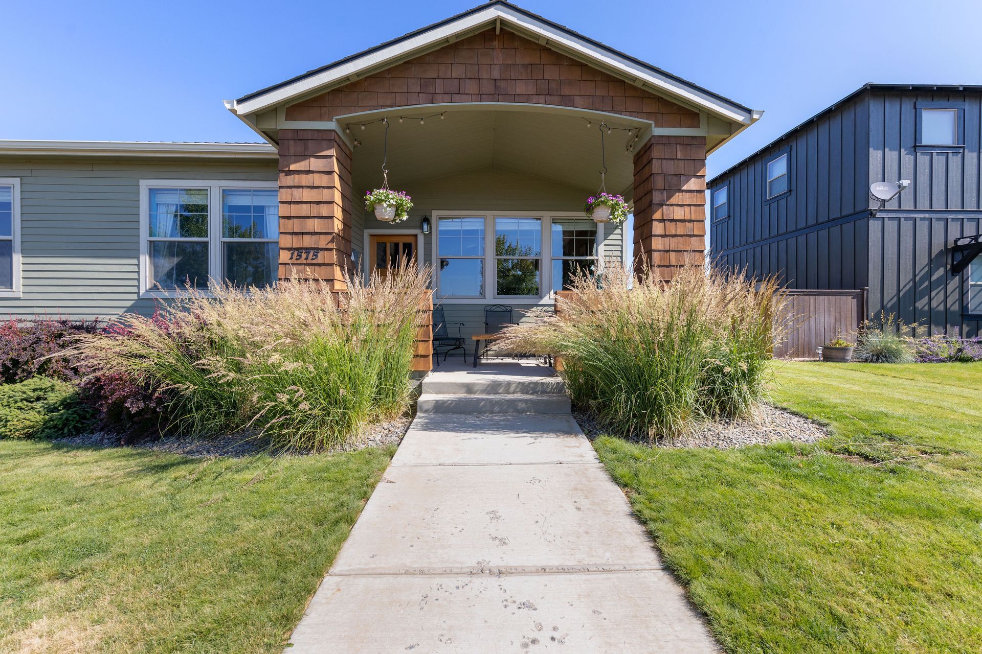 A walkway leads to the front porch of a bungalow-style home with tan siding and brown brick pillars, flanked by tall grass.