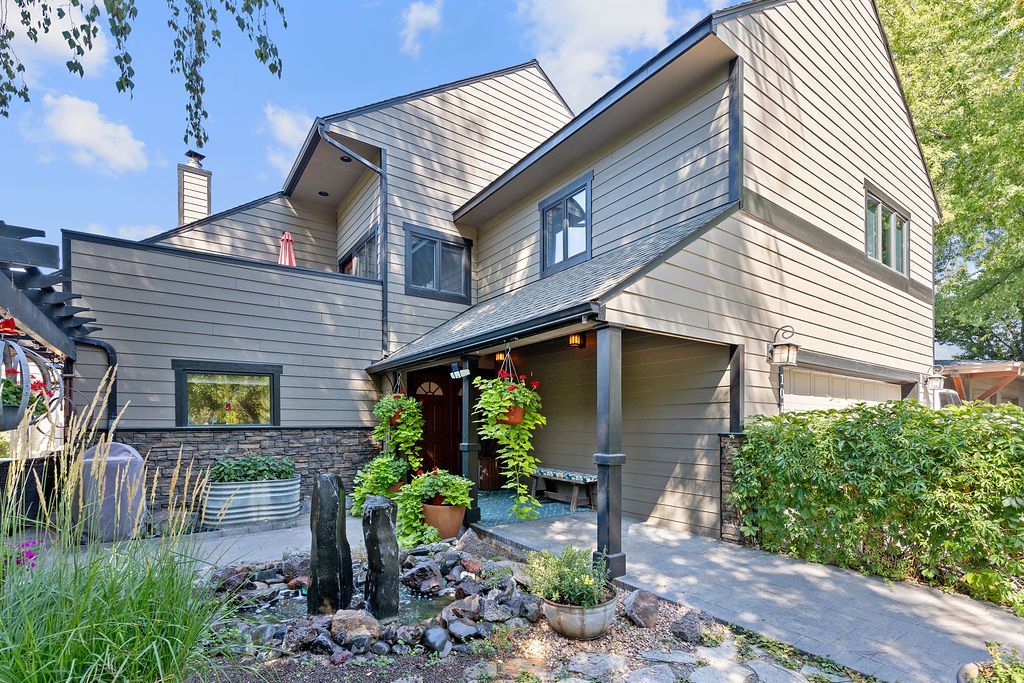 A two-story tan house with a dark roof and trim, featuring a front patio with stone landscaping and potted plants.