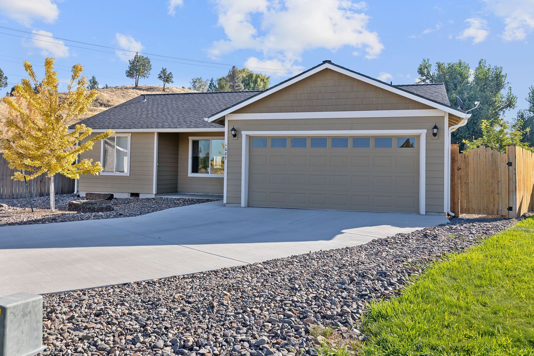 A single-story tan house with a dark roof and a two-car garage, featuring a concrete driveway and landscaped front yard.