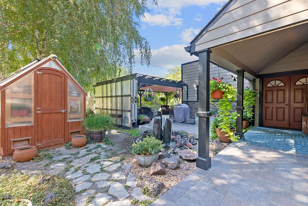 A backyard garden with a wooden shed, a greenhouse, stone pathways, and a covered patio area with a wooden door.