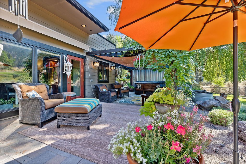 A sunny outdoor patio with wicker seating, a footstool, flowering plants in the foreground, and an orange umbrella.