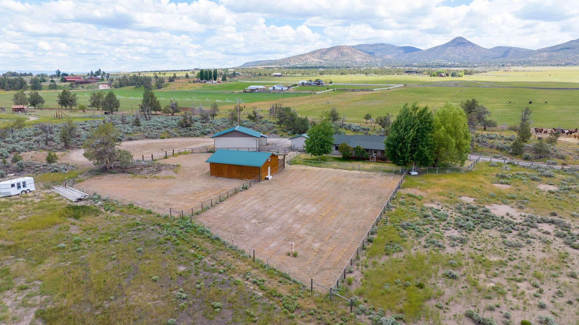 An aerial view of a wooden cabin with a green roof, fenced yard, and RV, set in a rural landscape with distant mountains.