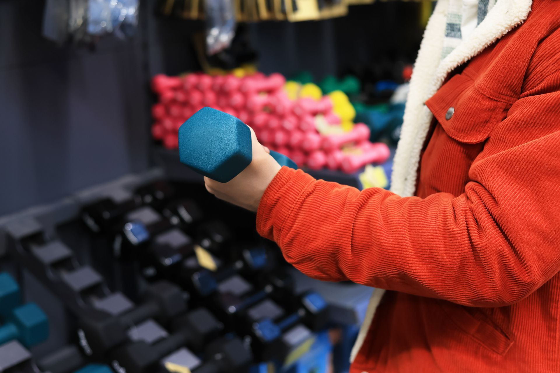 Close-up of a person wearing a red jacket as they hold a blue dumbbell inside a fitness shop. Close-up of a person wearing a red jacket as they hold a blue dumbbell inside a fitness shop.