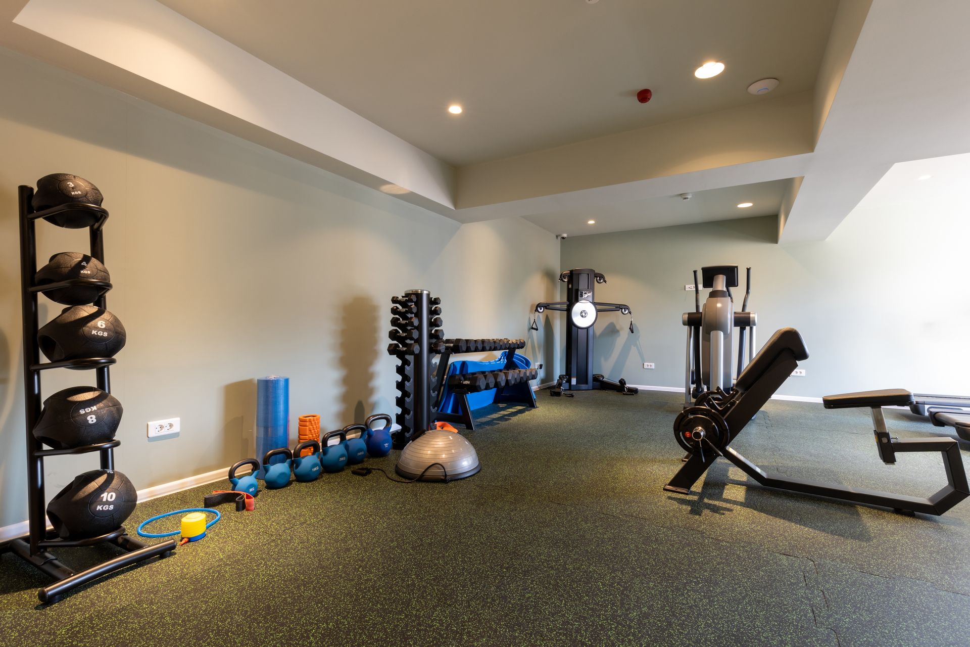A home gym equipment room with kettlebell rack, cable machine, incline bench, and rubber flooring.