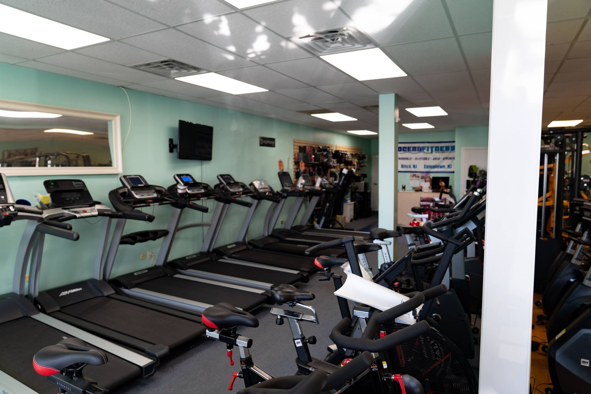 A gym interior featuring a row of treadmills against a light green wall and stationary bikes in the foreground.