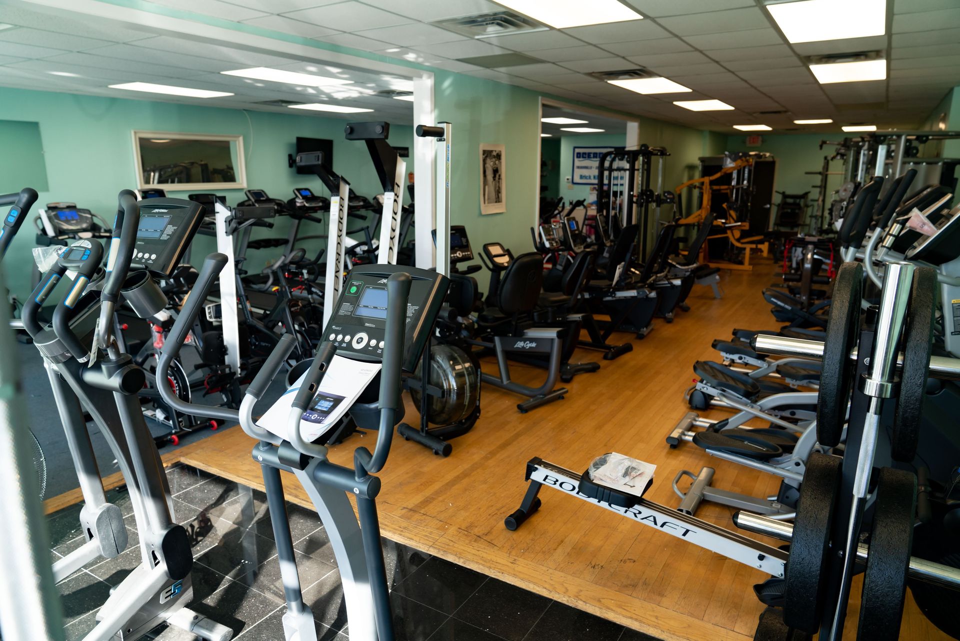 A gym featuring multiple rows of exercise machines, including ellipticals and stationary bikes on a wood-look floor.
